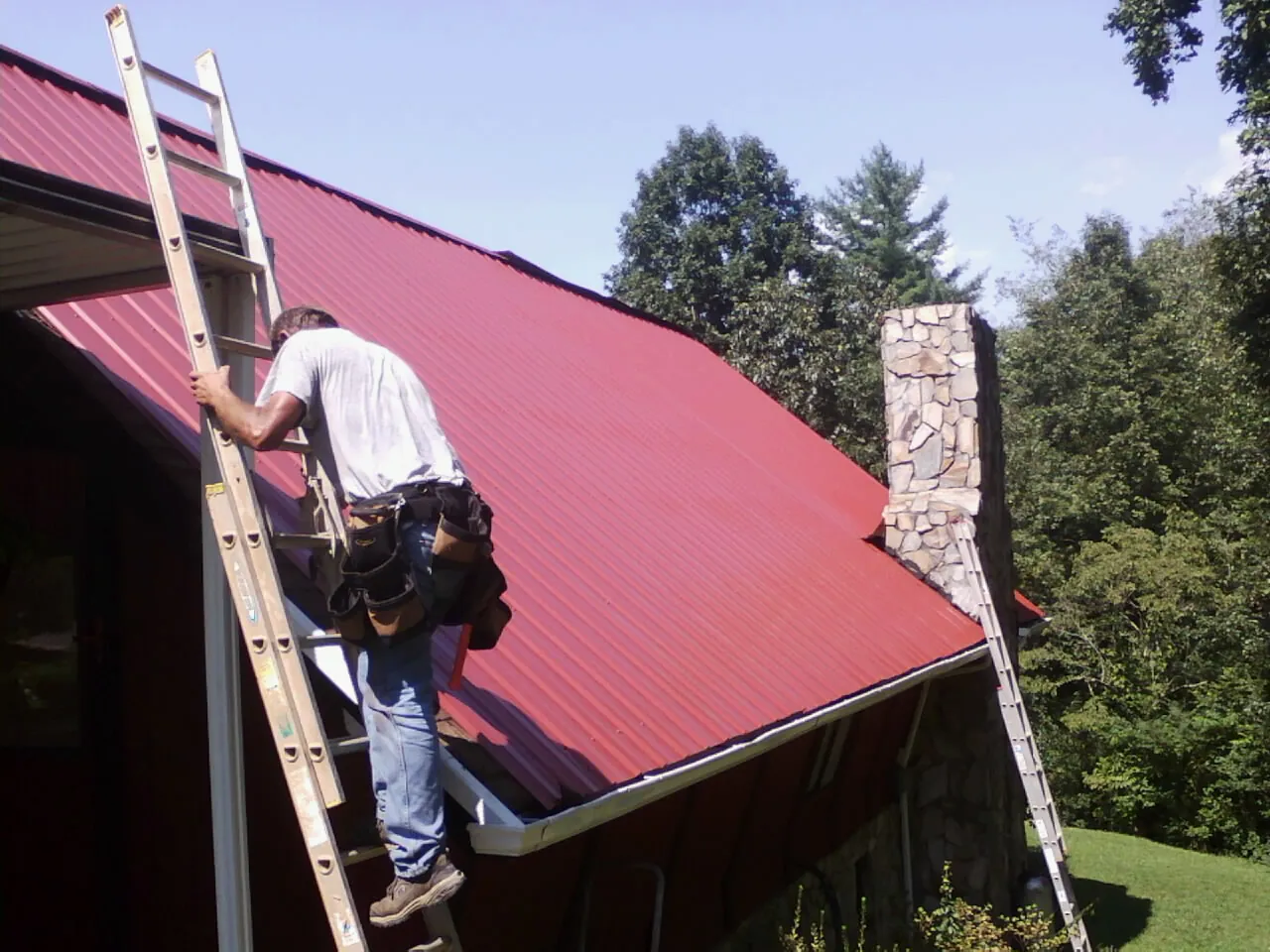 Roofer on a ladder beside a red metal roof and stone chimney — Soffit & Fascia Repair by Roofing Brothers in Grenloch